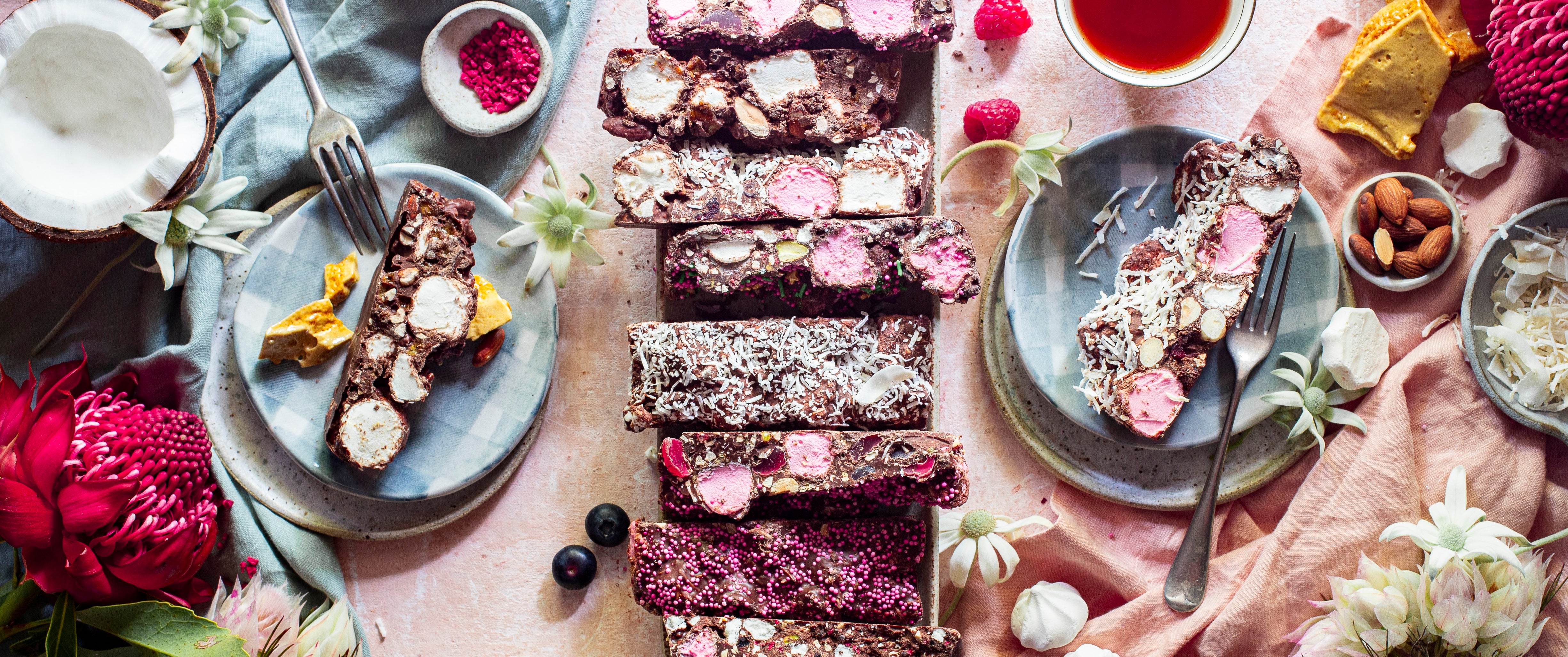 a light pink linen tablecloth is scattered with blocks of different flavoured chocolate rocky road, some on blue and white plates with dessert forks, and scattered with the local ingredients that go into the recipes, and there are some native flowers as decoration as well