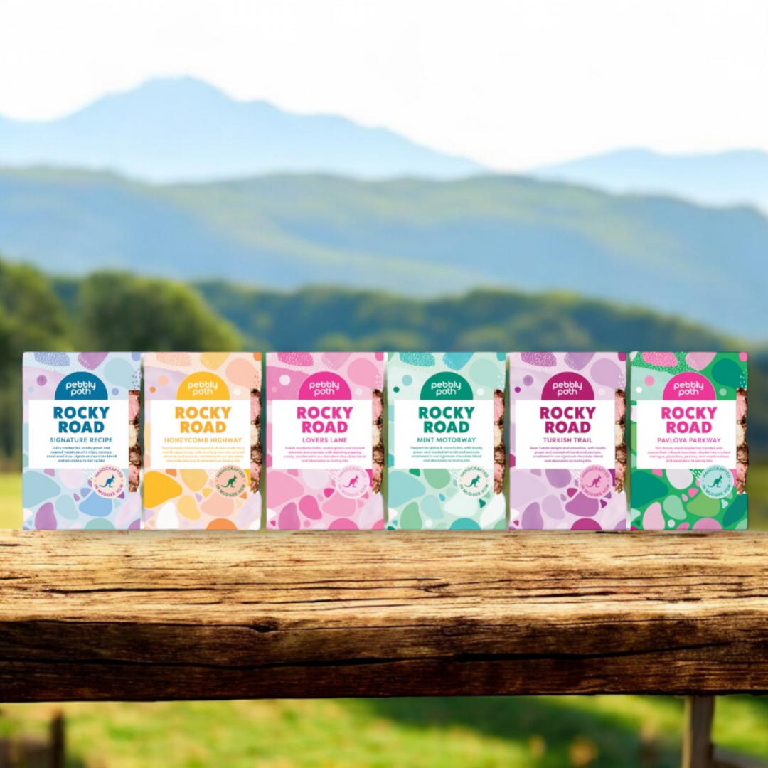 Assorted flavours of Rocky Road in bright, dopamine-inducing, 100% plastic-free packaging, on a rustic wooden table, with a stunning view of the bluey-green rolling hills of Mudgee NSW.