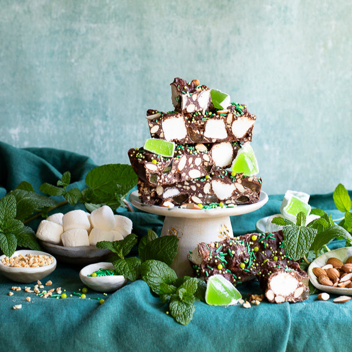 Dessert stand with white vanilla marshmallows and green mint flavoured jellies on a teal tablecloth against a textured blue wall, surrounded in mint leaves.