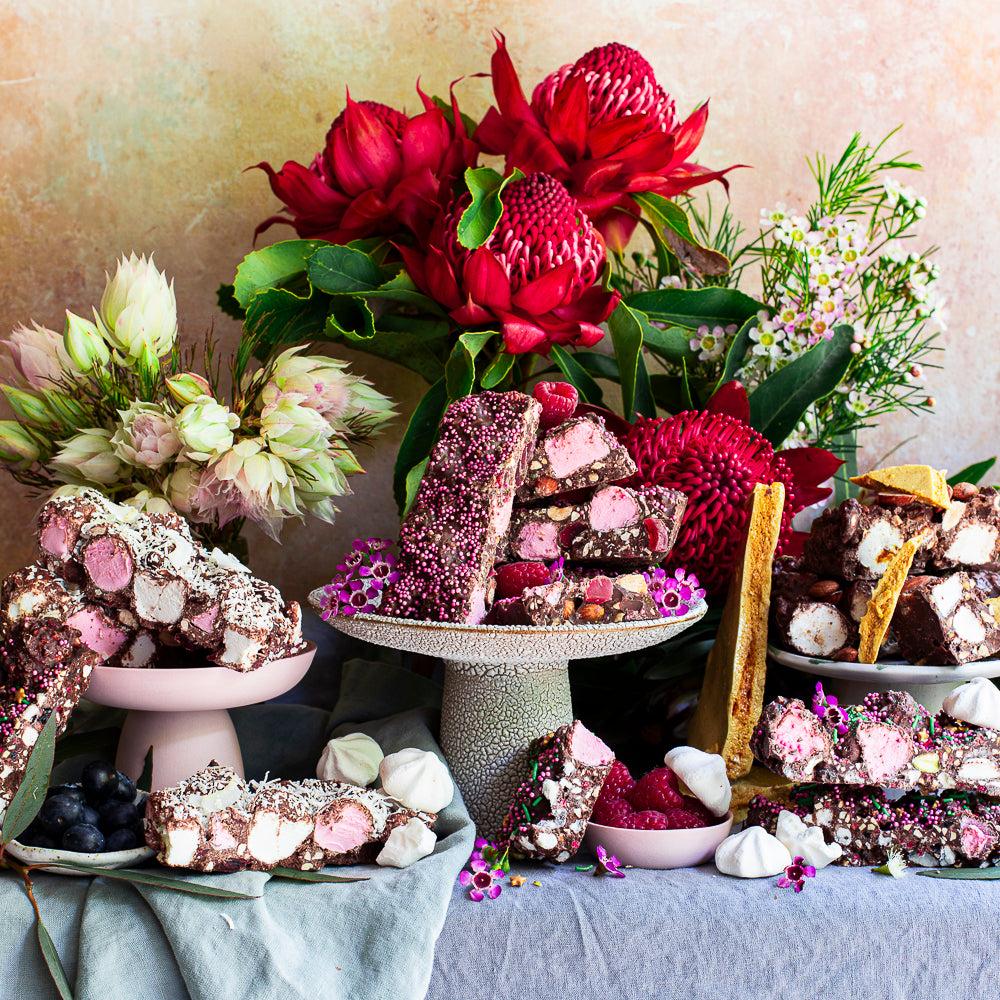 large chunks and blocks of different flavoured chocolate rocky roads are perched on a table and on plates and elevated cake platters.  the table has a rustic blue linen tablecloth, , and there are lots of native flowers scattered in the background. there are bowls of blueberries, raspberries, homemade honeycomb shards, meringues and other rocky road ingredients on the table as decoration.
