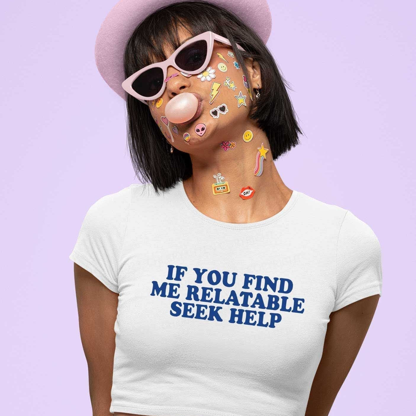 Person wearing a white crop top printed with the phrase “If you find me relatable seek help,” pink hat, pink sunglasses, and colourful stickers on their face, blowing bubble gum against a pastel purple background