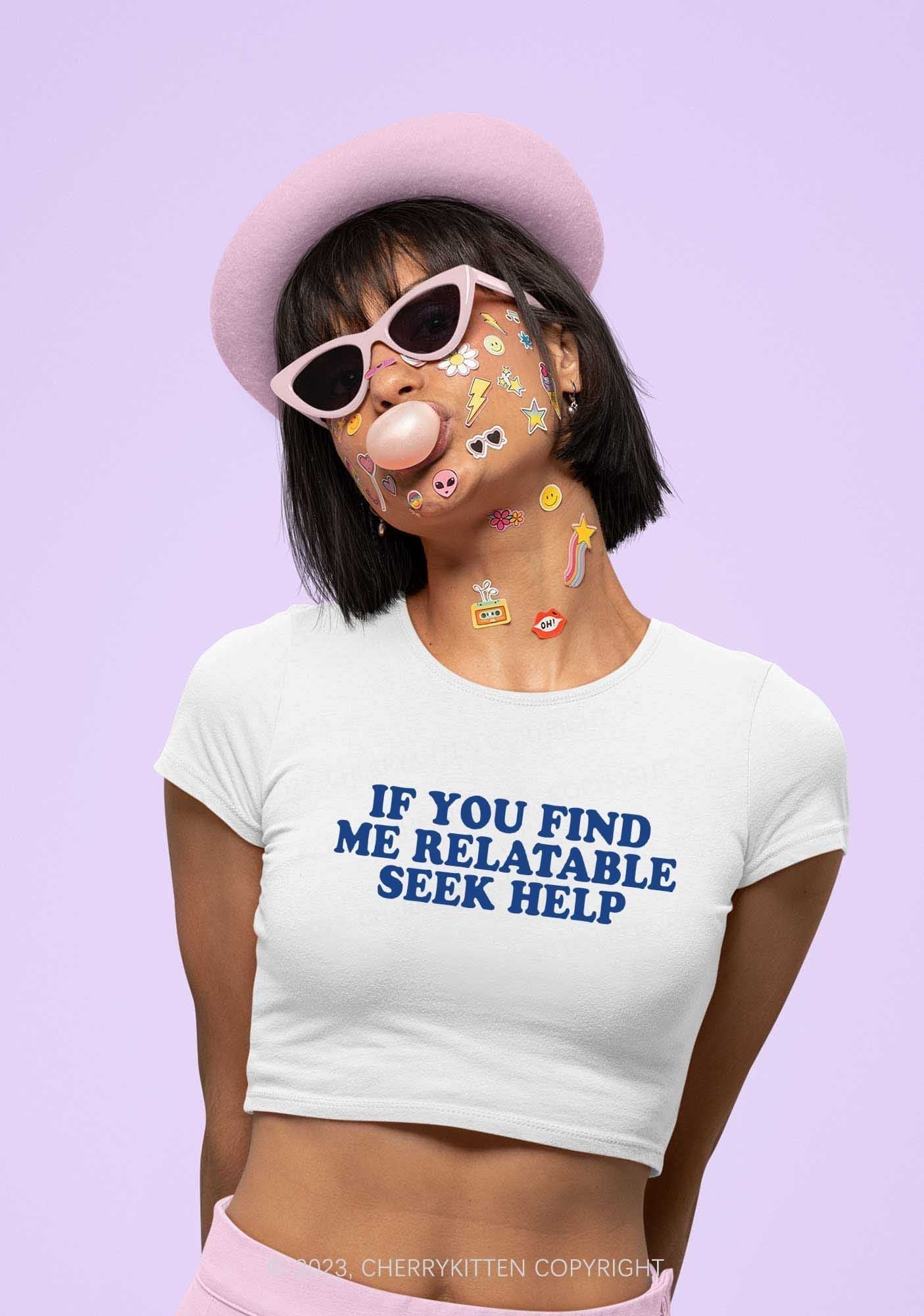 Person wearing a white crop top printed with the phrase “If you find me relatable seek help,” pink hat, pink sunglasses, and colourful stickers on their face, blowing bubble gum against a pastel purple background