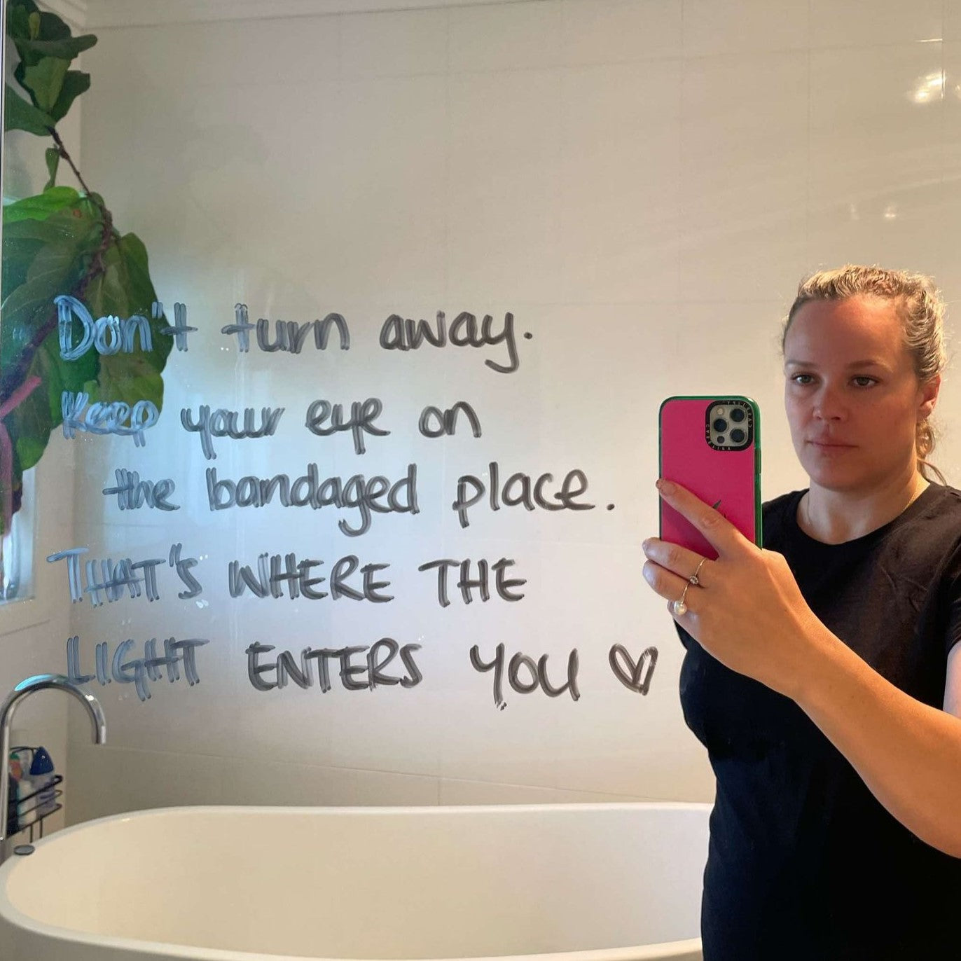 tash takes a mirror selfie of her bathroom mirror, with some words of affirmation written on the mirror. a wilted fiddle leaf fig struggles for life in the background.