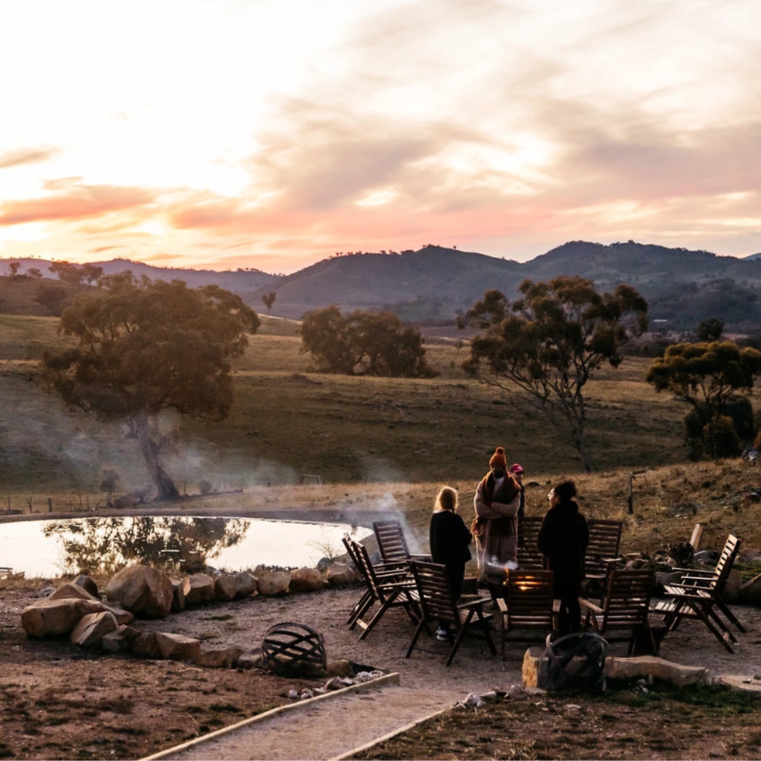 People rugged up around a firepit at sunset at Glenayr Farm, Mudgee, overlooking a dam and rolling hills in winter