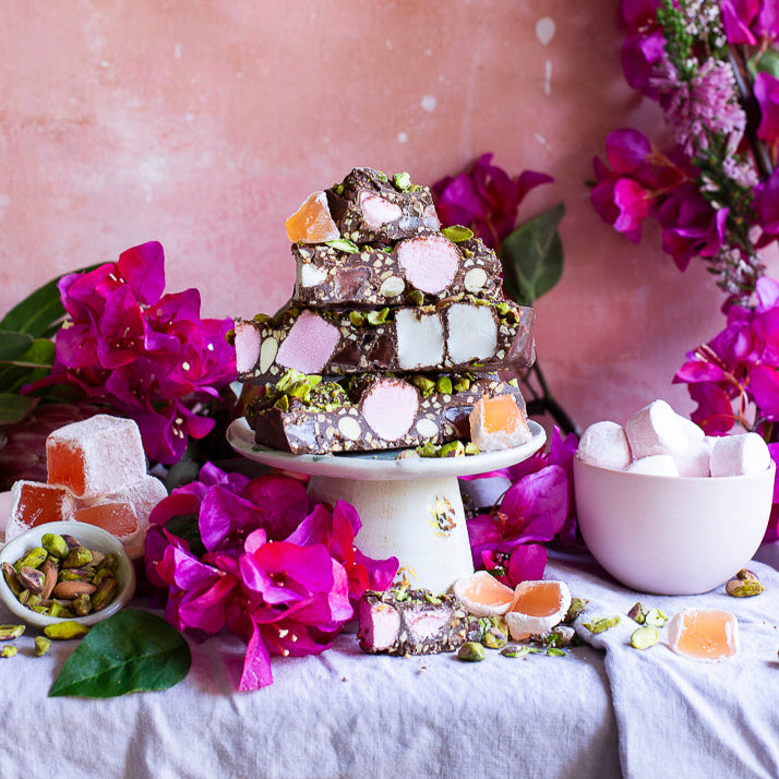 Pebbly Path Turkish Trail Rocky Road slices stacked on a pedestal with pistachios, Turkish delight and pink florals, styled as a decorative dessert display for sharing or entertaining.