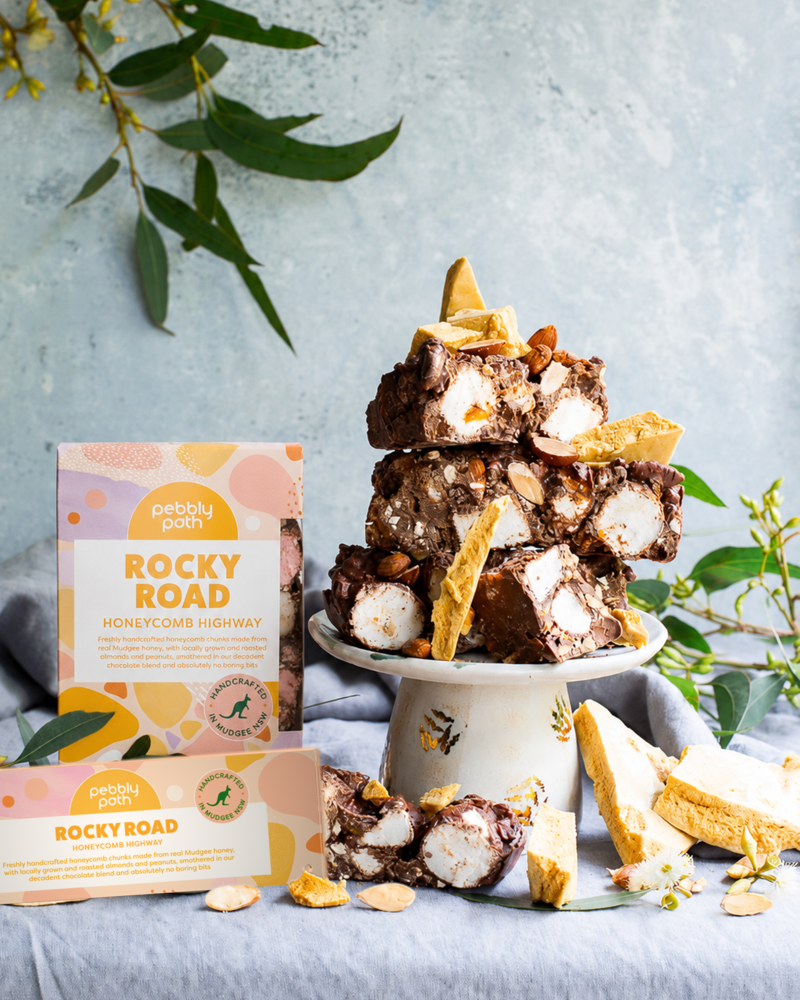 Pebbly Path Honeycomb Highway Rocky Road 200g and 100g boxes displayed with chunky rocky road pieces, honeycomb shards, marshmallows, almonds, and styled native greenery.