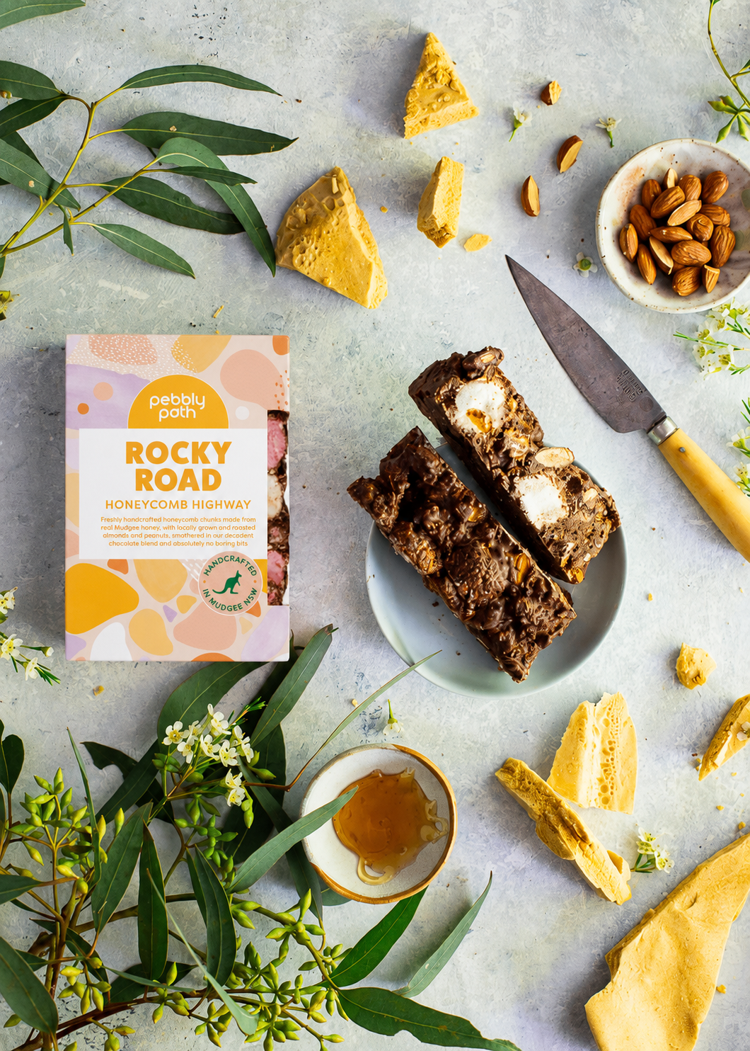 Pebbly Path Honeycomb Highway Rocky Road 200g box styled with sliced rocky road bars, honeycomb pieces, almonds, honey drizzle, and native greenery on a textured tabletop.