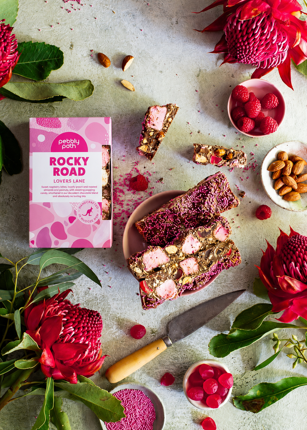 Pebbly Path Lovers Lane Rocky Road 200g box styled with sliced raspberry rocky road bars, pink marshmallows, almonds, raspberry lollies, fresh raspberries, and native flowers on a textured tabletop.