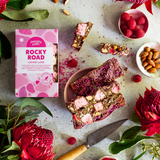Pebbly Path Lovers Lane Rocky Road 200g box styled with sliced raspberry rocky road bars, pink marshmallows, almonds, raspberry lollies, fresh raspberries, and native flowers on a textured tabletop.