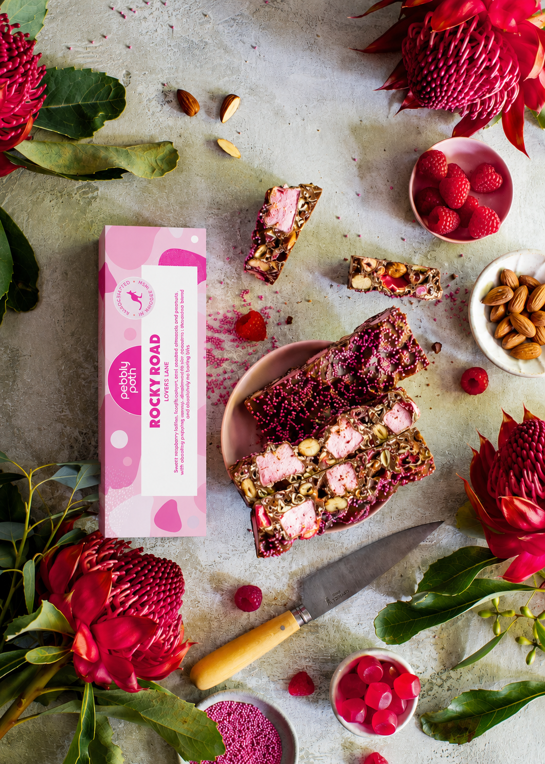 Pebbly Path Lovers Lane Rocky Road 100g box styled with sliced raspberry rocky road bars, pink marshmallows, almonds, raspberry lollies, fresh raspberries, and native flowers on a textured tabletop.