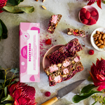 Pebbly Path Lovers Lane Rocky Road 100g box styled with sliced raspberry rocky road bars, pink marshmallows, almonds, raspberry lollies, fresh raspberries, and native flowers on a textured tabletop.