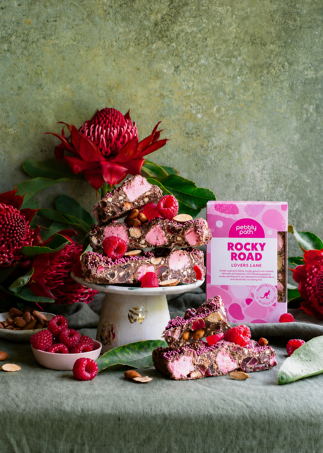 Pebbly Path Lovers Lane Rocky Road 200g box displayed with raspberry rocky road pieces, pink marshmallows, almonds, fresh raspberries, and native flowers on a styled dessert table.