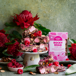 Pebbly Path Lovers Lane Rocky Road 200g box displayed with raspberry rocky road pieces, pink marshmallows, almonds, fresh raspberries, and native flowers on a styled dessert table.
