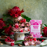Pebbly Path Lovers Lane Rocky Road 200g box displayed with raspberry rocky road pieces, pink marshmallows, almonds, fresh raspberries, and native flowers on a styled dessert table.