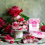 Pebbly Path Lovers Lane Rocky Road 200g and 100g boxes displayed with raspberry rocky road pieces, marshmallows, almonds, fresh raspberries, and native flowers in a styled dessert setting.
