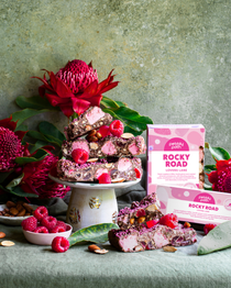 Pebbly Path Lovers Lane Rocky Road 200g and 100g boxes displayed with raspberry rocky road pieces, marshmallows, almonds, fresh raspberries, and native flowers in a styled dessert setting.