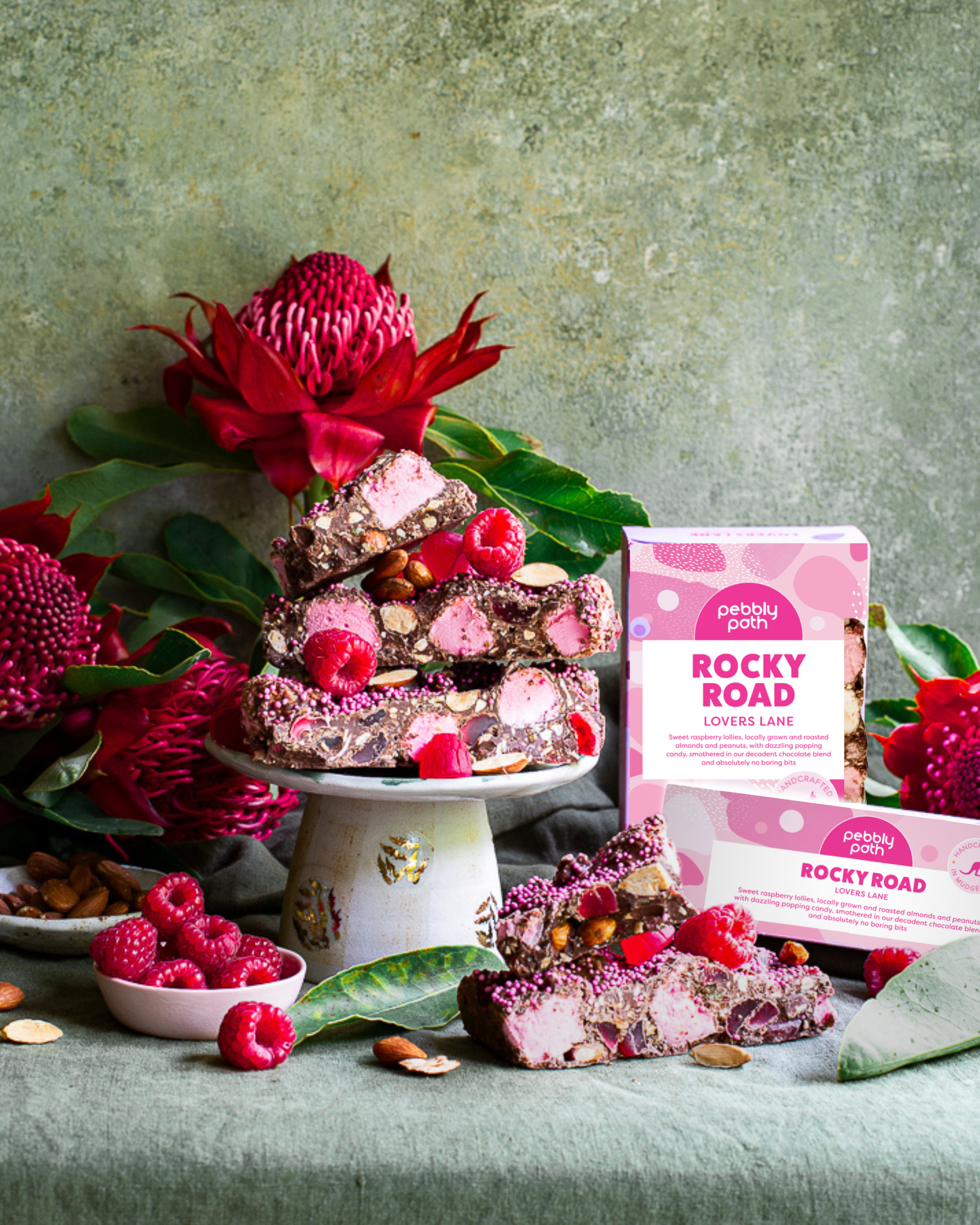 Pebbly Path Lovers Lane Rocky Road 200g and 100g boxes displayed with raspberry rocky road pieces, marshmallows, almonds, fresh raspberries, and native flowers in a styled dessert setting.