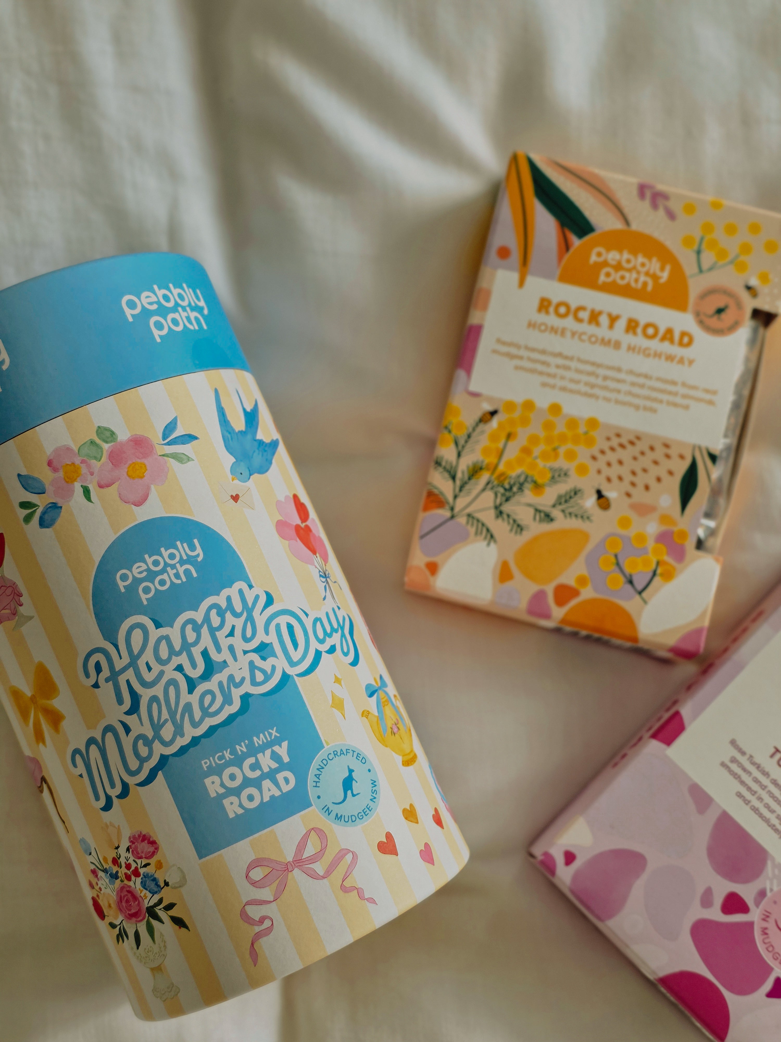 Pebbly Path Lovers Lane Valentine’s Day rocky road shown stacked on a cake stand with pink marshmallow, raspberry pieces, almonds and popping candy, styled with fresh raspberries and flowers as a chocolate gift.