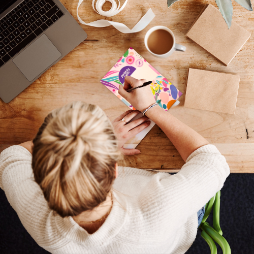 A person writing a handwritten gift card beside Pebbly Path gift packaging on a desk, capturing a thoughtful chocolate gifting moment.