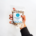 Pebbly Path Hazel-Coconut Ice Cream Crumble in a clear resealable stand-up pouch, held in one hand with the front product label visible, showing the chocolate, coconut, and hazelnut crumble blend through the packaging.