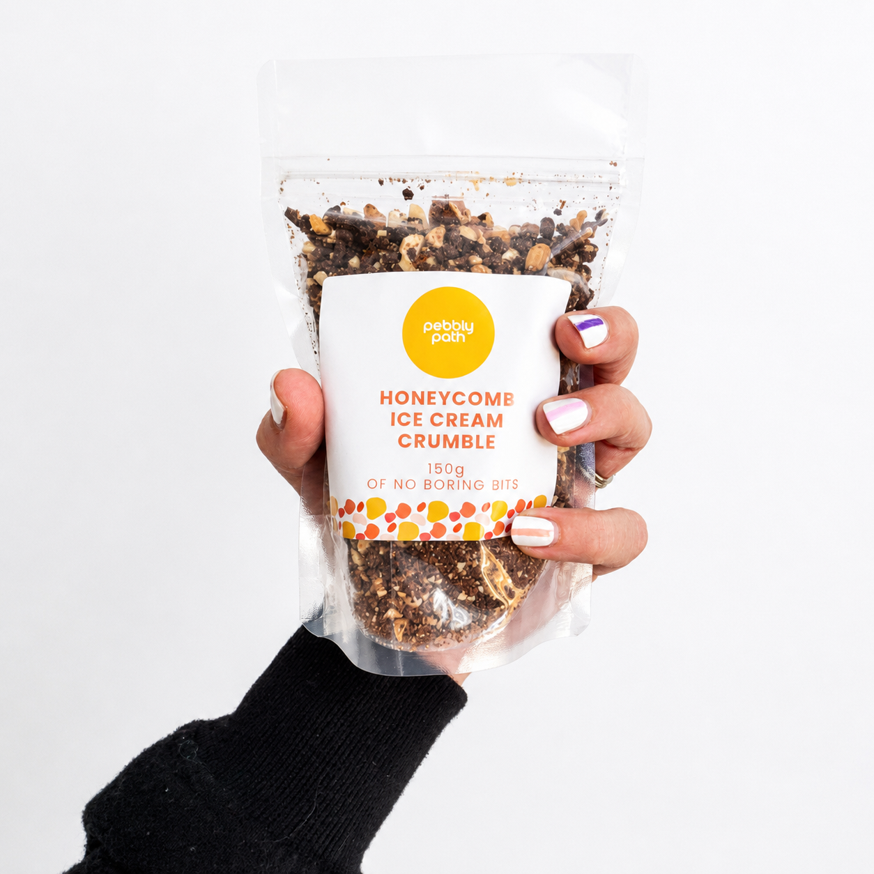 Pebbly Path Honeycomb Ice Cream Crumble in a clear resealable stand-up pouch, held in one hand with the front product label visible, showing the chocolate and honeycomb crumble blend through the packaging.