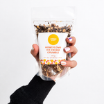Pebbly Path Honeycomb Ice Cream Crumble in a clear resealable stand-up pouch, held in one hand with the front product label visible, showing the chocolate and honeycomb crumble blend through the packaging.
