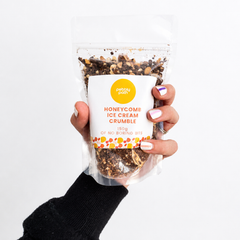 Pebbly Path Honeycomb Ice Cream Crumble in a clear resealable stand-up pouch, held in one hand with the front product label visible, showing the chocolate and honeycomb crumble blend through the packaging.