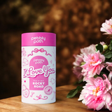 Pebbly Path Pick n Mix “I Love You” rocky road cylinder, an Australian handmade chocolate gift styled on a wooden table beside pink flowers, ideal for romantic gifting.