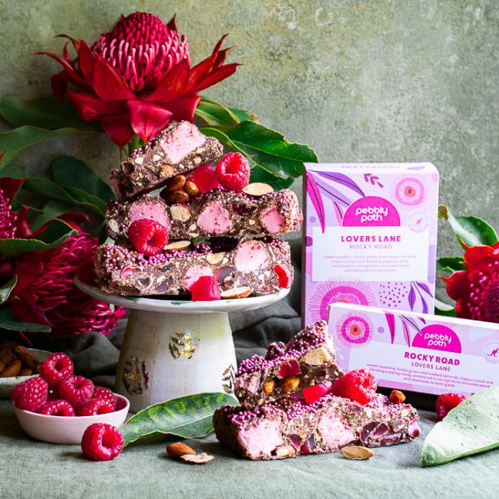 Pebbly Path Lovers Lane Rocky Road displayed with raspberry pieces, pink marshmallows and roasted almonds, styled with native flowers and branded packaging in a premium product display.