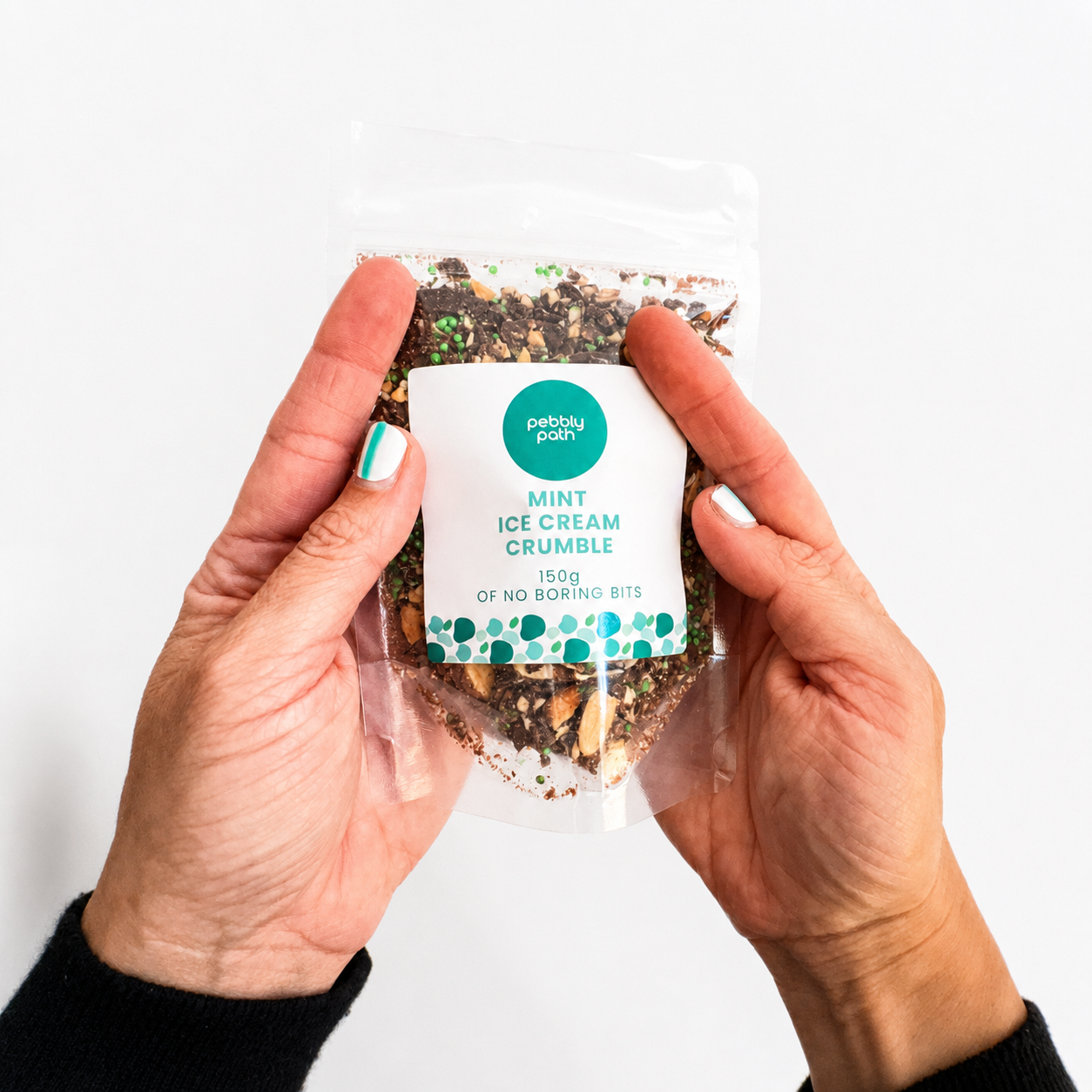 Pebbly Path Mint Ice Cream Crumble in a clear resealable stand-up pouch, held in both hands with the front product label visible, showing the chocolate, mint pieces, and crunchy crumble blend through the packaging.