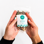 Pebbly Path Mint Ice Cream Crumble in a clear resealable stand-up pouch, held in both hands with the front product label visible, showing the chocolate, mint pieces, and crunchy crumble blend through the packaging.