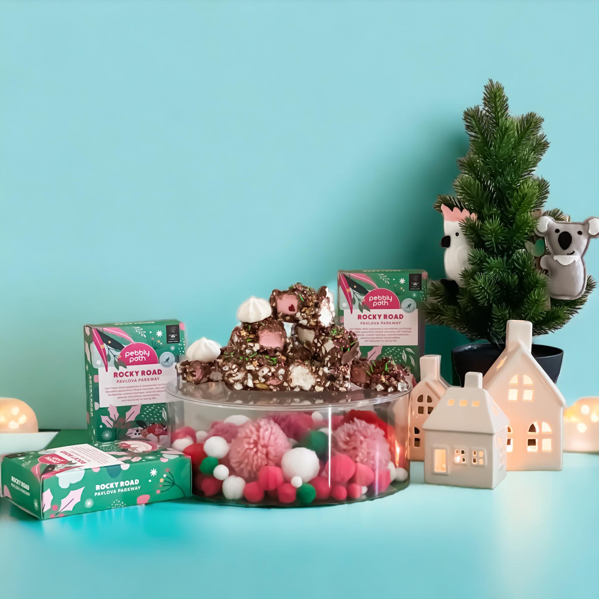 Pebbly Path Pavlova Parkway Rocky Road Christmas display with stacked rocky road pieces, festive packaging, meringue and holiday decorations beside illuminated ceramic houses and a mini Christmas tree