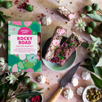 Pebbly Path Pavlova Parkway 200g rocky road box beside sliced chocolate rocky road pieces on a plate, with raspberries, pistachios and mini meringues styled around the scene.