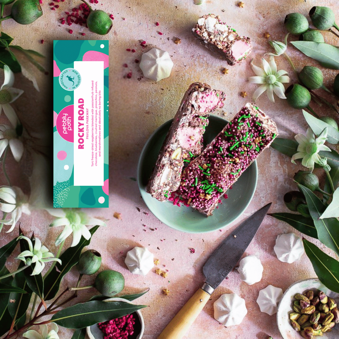Pebbly Path Pavlova Parkway 100g Rocky Road bars with colourful sprinkles, pink marshmallows, pistachios and meringue beside product packaging in a styled flat lay display