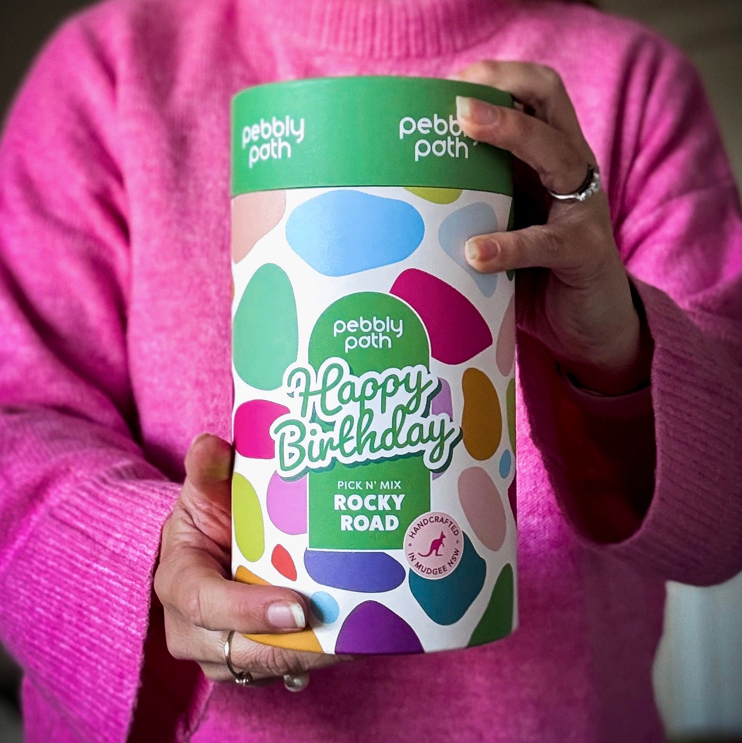 Pebbly Path Pick n Mix Rocky Road Cylinder with a Happy Birthday design, held in both hands by a person wearing a bright pink sweater, showcasing the colourful gift cylinder packaging for birthday gifting.
