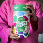 Pebbly Path Pick n Mix Rocky Road Cylinder with a Happy Birthday design, held in both hands by a person wearing a bright pink sweater, showcasing the colourful gift cylinder packaging for birthday gifting.