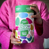Pebbly Path Pick n Mix Rocky Road Cylinder with a Happy Birthday design, held in both hands by a person wearing a bright pink sweater, showcasing the colourful gift cylinder packaging for birthday gifting.