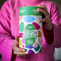 Pebbly Path Pick n Mix Rocky Road Cylinder with a Happy Birthday design, held in both hands by a person wearing a bright pink sweater, showcasing the colourful gift cylinder packaging for birthday gifting.