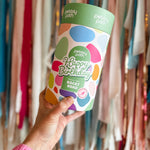 Pebbly Path Pick n Mix Rocky Road Cylinder with a Happy Birthday design, held in hand against a backdrop of colourful party streamers, showcasing a festive birthday gift option.