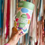 Pebbly Path Pick n Mix Rocky Road Cylinder with a Happy Birthday design, held in hand against a backdrop of colourful party streamers, showcasing a festive birthday gift option.