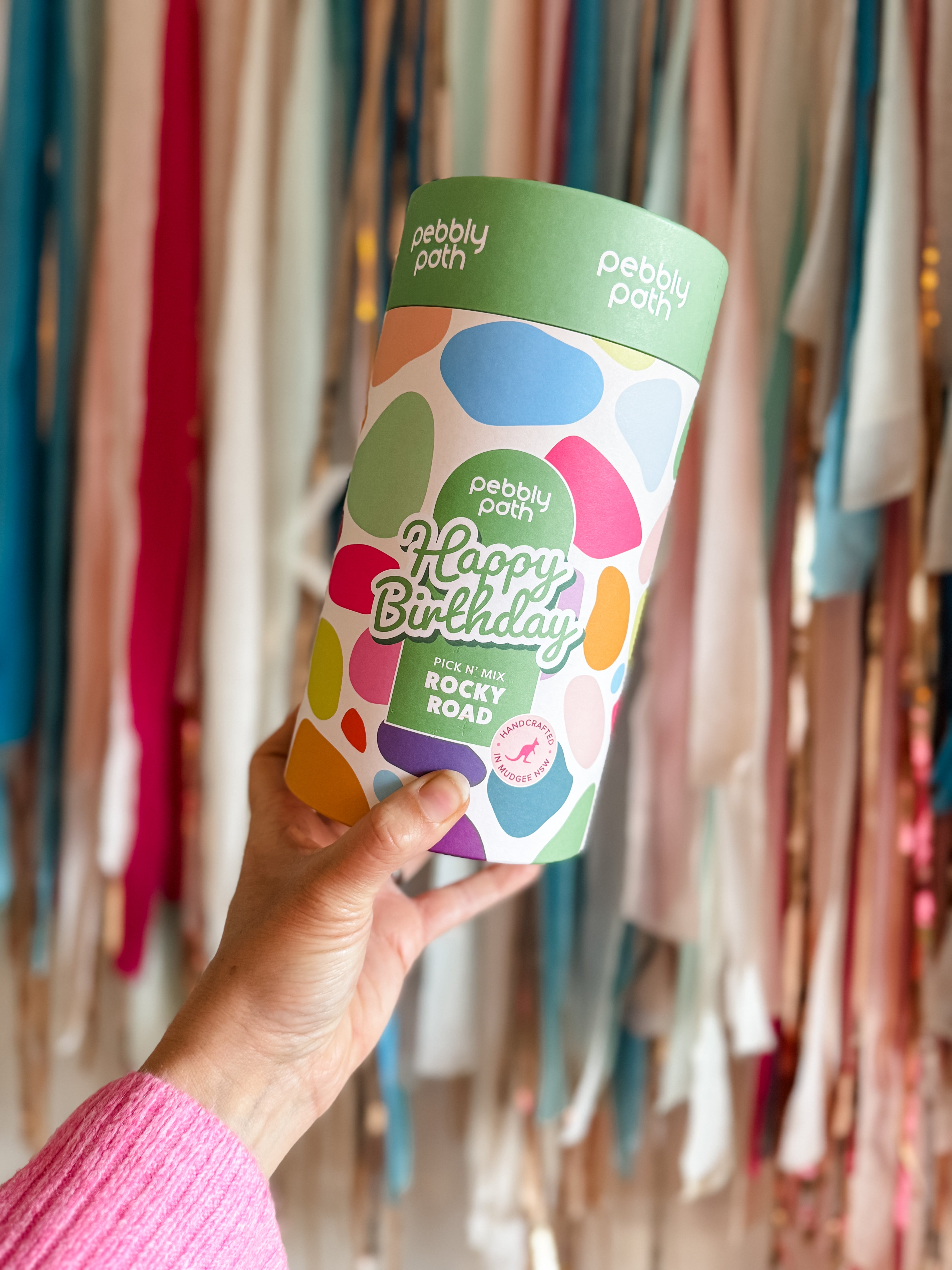 Pebbly Path Pick n Mix Rocky Road Cylinder with a Happy Birthday design, held in hand against a backdrop of colourful party streamers, showcasing a festive birthday gift option.