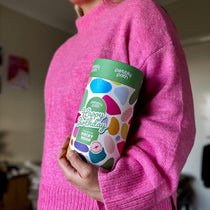 Pebbly Path Pick n Mix Rocky Road Cylinder with a Happy Birthday design, held under one arm by a person wearing a bright pink sweater, highlighting the gift cylinder’s size and colourful packaging.
