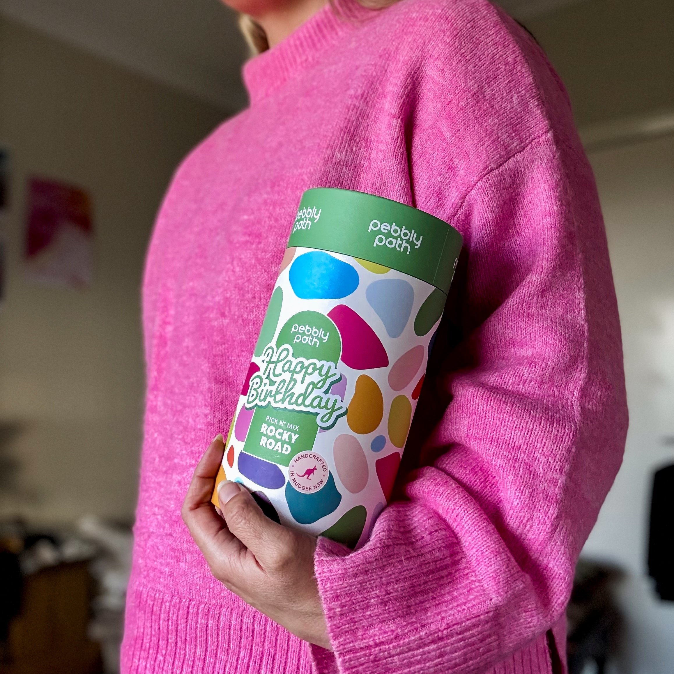 Pebbly Path Pick n Mix Rocky Road Cylinder with a Happy Birthday design, held under one arm by a person wearing a bright pink sweater, highlighting the gift cylinder’s size and colourful packaging.