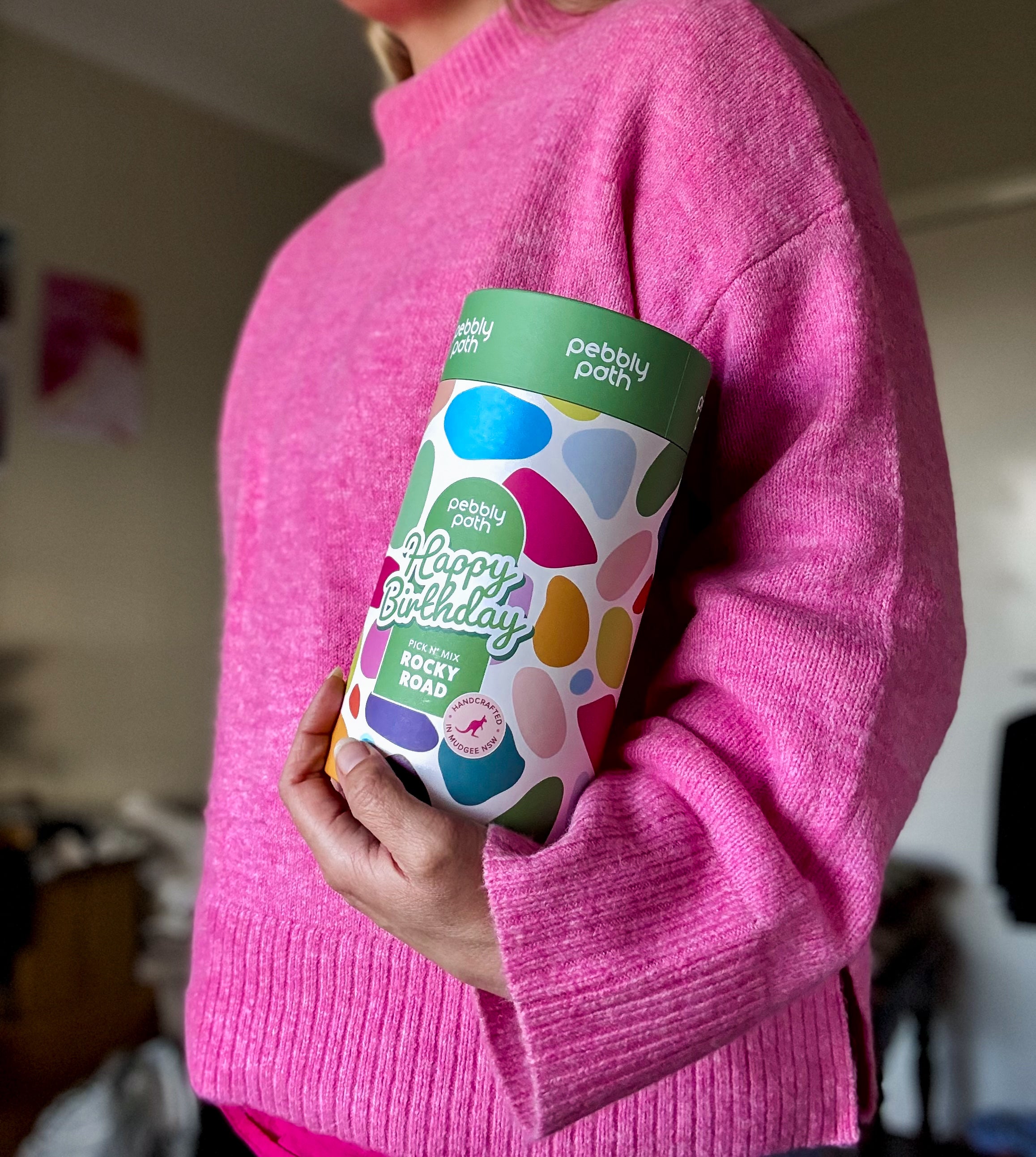 Pebbly Path Pick n Mix Rocky Road Cylinder with a Happy Birthday design, held under one arm by a person wearing a bright pink sweater, highlighting the gift cylinder’s size and colourful packaging.