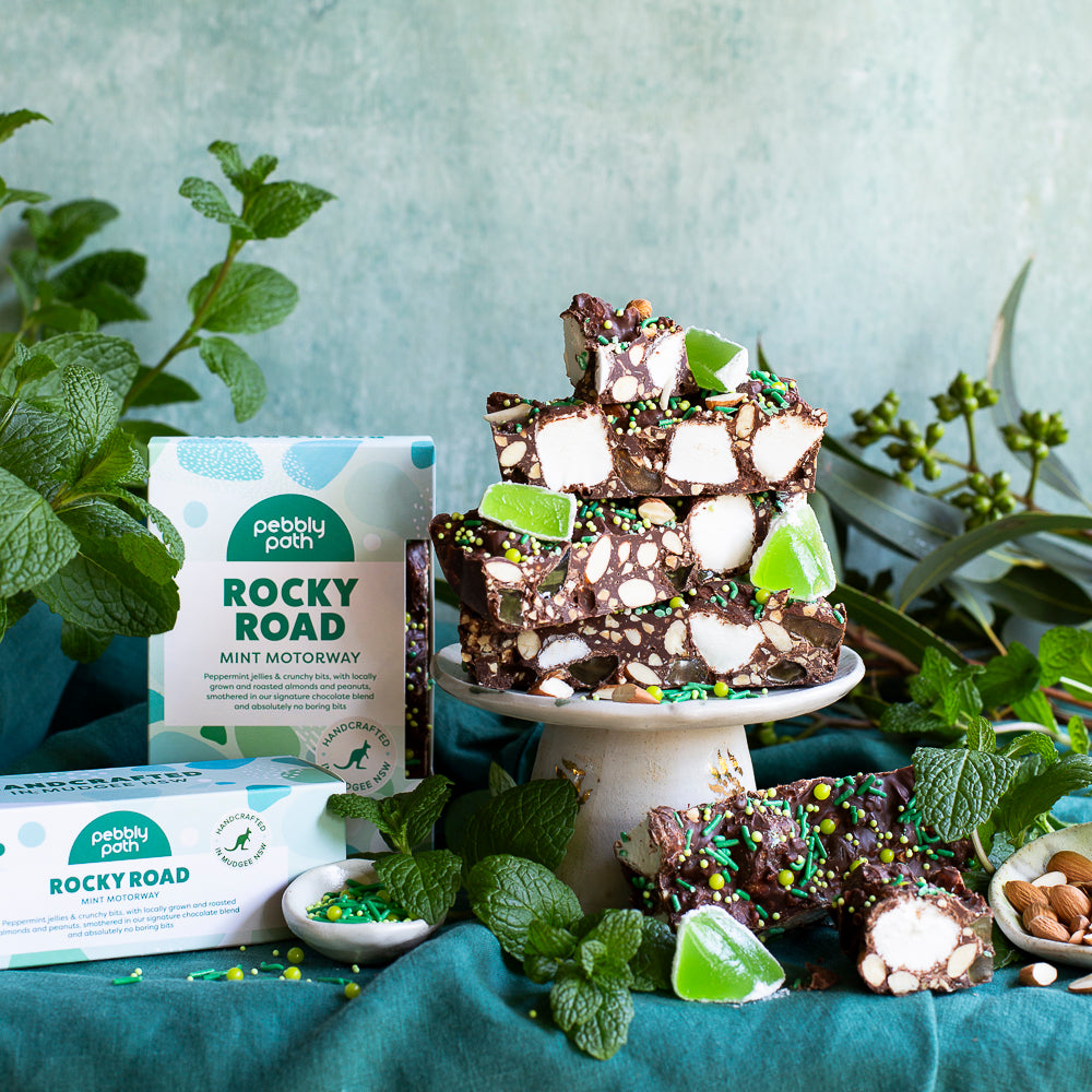 Pebbly Path Mint Motorway Rocky Road grouped 100g and 200g boxes displayed beside stacked rocky road slices on a pedestal with fresh mint leaves and green jelly pieces, styled as a handcrafted chocolate gift or dessert centrepiece.