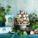 Pebbly Path Mint Motorway Rocky Road grouped 100g and 200g boxes displayed beside stacked rocky road slices on a pedestal with fresh mint leaves and green jelly pieces, styled as a handcrafted chocolate gift or dessert centrepiece.