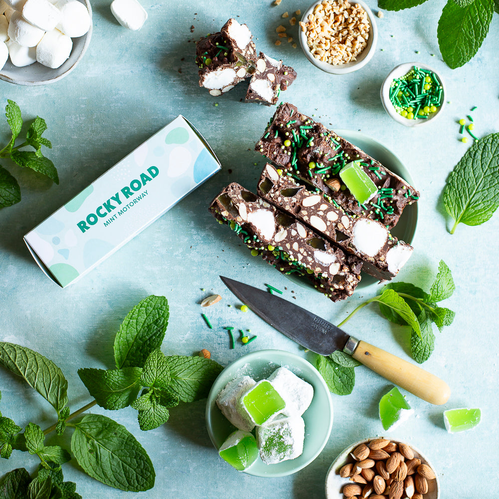 Pebbly Path Mint Motorway Rocky Road 100g box shown alongside sliced rocky road bars with visible marshmallow, nuts and mint pieces, arranged as a flat lay for tasting, serving or dessert styling.