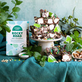 Pebbly Path Mint Motorway Rocky Road 200g box shown with stacked slices, mint leaves and scattered ingredients on a pedestal, presented as a fresh chocolate dessert option for sharing or entertaining.