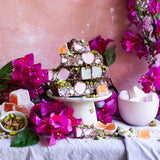 Pebbly Path Turkish Trail Rocky Road slices stacked on a pedestal with pistachios, Turkish delight and pink florals, styled as a decorative dessert display for sharing or entertaining.