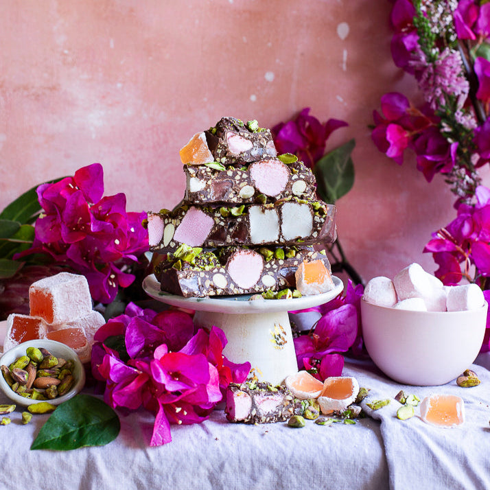 Pebbly Path Turkish Trail Rocky Road slices stacked on a pedestal with pistachios, Turkish delight and pink florals, styled as a decorative dessert display for sharing or entertaining.